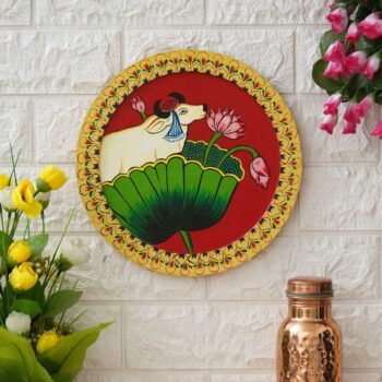 This hand-painted pichwai depicts a sacred cow sitting on a lotus flower. The cow is a symbol of fertility, abundance, and motherhood in Hinduism, and the lotus flower is a symbol of purity and enlightenment. The painting is made with natural pigments on cotton cloth, and it is framed in a traditional wooden frame. This unique piece of art would be a beautiful addition to any home.
